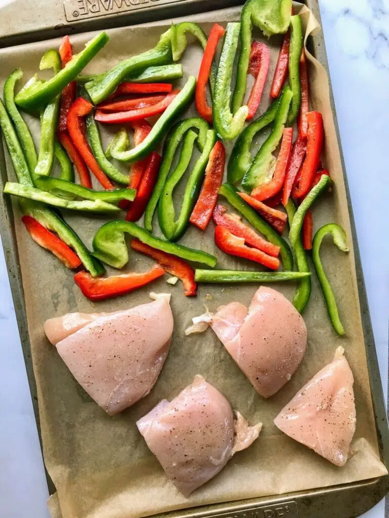 raw red and green bell peppers and raw chicken breast on a baking sheet.